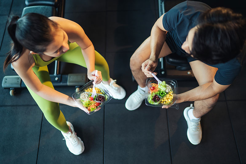 Two athletes eating prepared high protein meals on the gym floor